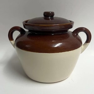 May include: A two-handled ceramic bean pot with a glossy dark brown glazed upper section and a matte cream-colored lower body. The pot features a matching dark brown lid with a central knob handle and two curved side handles for lifting.