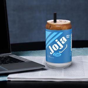 Könnte beinhalten: Ein Glas mit "Joja Cola"-Aufdruck und einem schwarzen Strohhalm. Das Glas hat ein blau-weißes Etikett. Das Getränk ist hellbraun. Der Text "Life's Better With Joja!" ist auf der Seite des Glases aufgedruckt.
