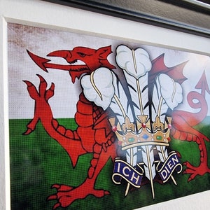 May include: Framed artwork of the Welsh flag, featuring a red dragon and the motto "Ich Dien." The artwork is framed in black with a white mount. The flag design includes a green and white background.