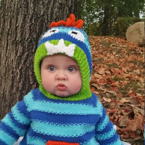 May include: A baby wearing a blue, green, and orange striped sweater with a matching monster hat. The hat has a white tooth and eyes. The baby is sitting on a white blanket.
