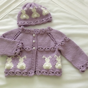 Puede incluir: Conjunto de cárdigan y gorro de bebé de punto lila. El cárdigan presenta diseños de conejitos blancos y botones de perlas. El gorro tiene diseños similares. El conjunto está hecho de hilo suave, ideal para un bebé.