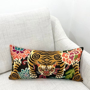 May include: A black and gold lumbar pillow with a tiger and floral design. The tiger is facing forward with its mouth open and its eyes are closed. The flowers are in shades of pink, red, and yellow.