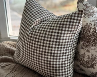 May include: Two decorative throw pillows. One pillow features a brown and white gingham pattern. The other pillow has a toile design in brown and white. The pillows are resting on a neutral-colored surface near a window.