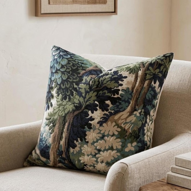 May include: A decorative throw pillow with a forest scene design. The pillow features a detailed pattern of trees and foliage in shades of green, blue, and brown. The pillow is placed on a neutral-colored armchair, suggesting a home decor theme.