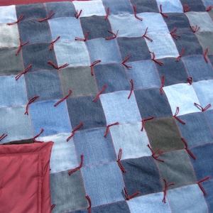 May include: A patchwork quilt made from squares of denim in various shades of blue and gray. The squares are joined with red thread and the quilt has a burgundy border. The quilt is a cozy, handmade item.