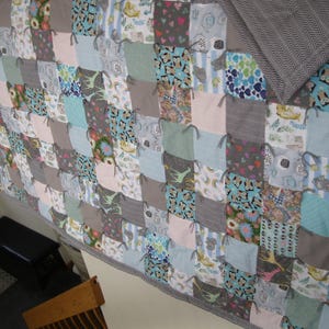 May include: A handmade patchwork quilt with a variety of patterned squares in shades of gray, blue, pink, and green. The quilt features a gray chevron patterned backing and is tied with gray yarn. The quilt is a cozy home decor item.