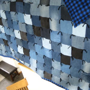 May include: A patchwork quilt constructed from square denim fabric pieces in various shades of blue and black. The squares are joined together with black fabric ties at each corner, and the quilt features a blue and black buffalo check border.