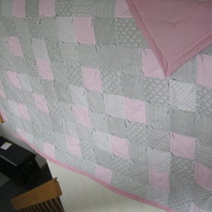 May include: A handmade rag quilt featuring a patchwork design of alternating solid light pink squares and various grey patterned squares, including chevron, lattice, and textured prints, finished with a solid light pink fabric border.