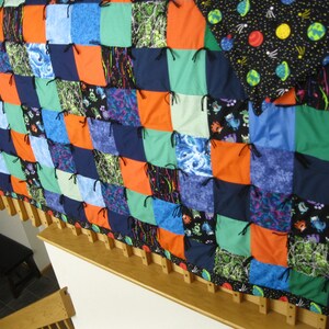 May include: A colorful patchwork quilt with squares in shades of blue, orange, green, and patterned fabrics. The quilt is secured with black ties and has a black border with a space-themed pattern. The quilt is displayed on a wooden frame.