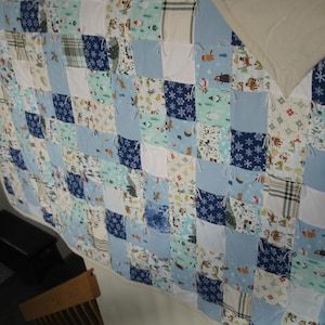 May include: A handmade patchwork quilt featuring a variety of fabrics in shades of blue, white, and cream. The quilt is composed of square patches with snowflake, plaid, and winter-themed patterns. The quilt is a cozy home decor item.
