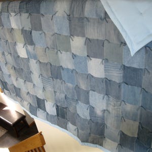 May include: A handmade quilt featuring a patchwork design of various shades of blue denim squares. The quilt has a light blue border and a light blue square in the upper right corner. The quilt is a cozy, handcrafted item.