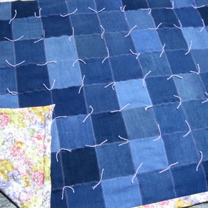 May include: A patchwork quilt constructed from square denim fabric pieces in various shades of blue. The squares are joined with zigzag stitching and secured with light pink yarn ties. One corner is folded back to reveal a floral print fabric lining.