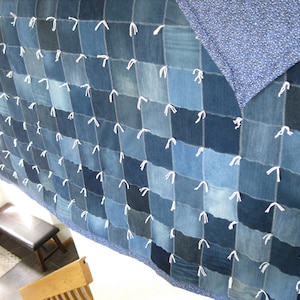 May include: A patchwork quilt constructed from square denim fabric pieces in various shades of blue. The squares are joined with white thread ties at each corner, and the quilt features a dark blue patterned fabric border and backing.