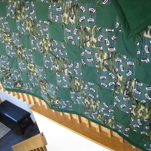May include: A handmade tie-fringe fleece blanket featuring a patchwork design of solid dark green squares, camouflage print squares, and squares with a pattern of white American footballs on a dark green background with grid lines.