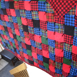 May include: A patchwork quilt featuring a variety of plaid patterns in red, black, blue, and green. The quilt is made of squares sewn together, with a red plaid border. The quilt is a cozy home decor item.