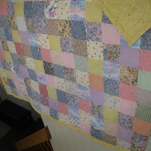 May include: A handmade patchwork quilt with a variety of pastel-colored fabrics. The quilt features squares of floral, polka dot, and solid patterns in shades of pink, blue, yellow, and lavender. The quilt is trimmed with a yellow border.