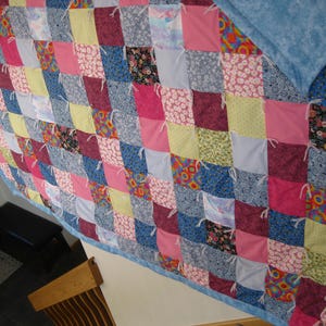 May include: A handmade quilt with a patchwork design. The quilt features a variety of fabrics in pink, blue, yellow, and floral patterns. The squares are tied together with white thread and have a blue border.