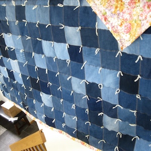 May include: A patchwork quilt constructed from square denim fabric pieces in various shades of blue, featuring white yarn ties at each corner intersection. The reverse side of the quilt displays a floral pattern with pink, yellow, and green colors.