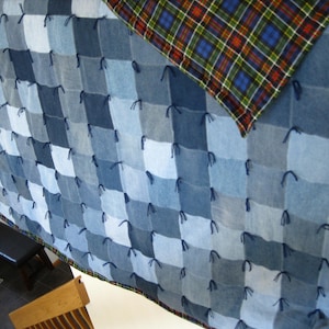 May include: A quilt constructed from square patches of blue denim fabric in varying shades, featuring dark blue yarn ties at each corner of the squares. The reverse side of the quilt displays a multi-colored plaid pattern in blue, green, and red.
