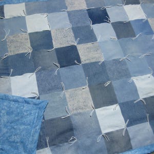 May include: A patchwork quilt constructed from square denim fabric pieces in various shades of blue, ranging from light to dark. The squares are joined with white yarn ties at each corner, and the quilt features a solid light blue fabric border.