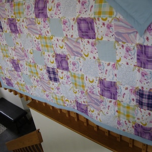May include: A patchwork quilt with a light blue border, featuring squares of various patterns and colors. The quilt includes squares of purple plaid, light blue, and floral designs with yellow accents. The quilt is displayed over a wooden structure.
