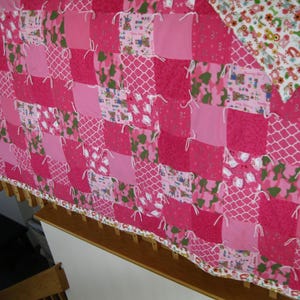 May include: A patchwork quilt in shades of pink, featuring a variety of patterns including solid squares, white lattice, and floral prints. The quilt is hand-tied with white yarn and has a floral border. A corner of the quilt has a white background with colorful flowers.