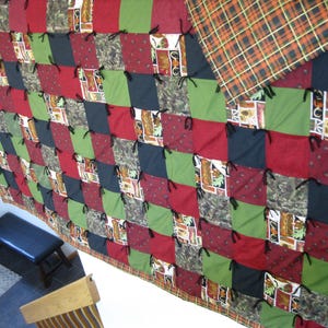 May include: A patchwork quilt featuring a grid of square fabric panels in shades of dark red, olive green, black, and a camouflage print. The squares are joined with black fabric ties at each corner, and the quilt has a plaid backing in orange and black.