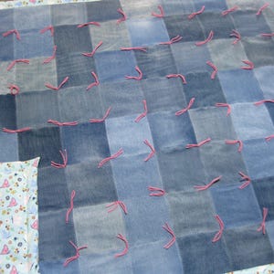 May include: A patchwork quilt constructed from various shades of blue denim fabric squares. The squares are joined with visible stitching and secured at the corners with small pink yarn ties. One corner reveals a light blue floral patterned fabric lining.