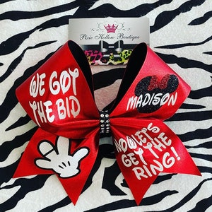 May include: Red cheer bow with white glitter lettering that says "We Got The Bid" on one side and "Now Let's Get The Ring!" on the other side. The bow also has a black Mickey Mouse head with glitter and the name "Madison" on it.