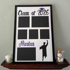 May include: A black framed graduation photo display with the text "Class of 2026" and "Nicolas" in glittery black and purple. The display has six black photo spaces and a silhouette of a graduate.