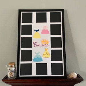 May include: A black picture frame with a white mat and 12 openings for photos. The frame has a princess theme with a crown and four princess dresses in various colors. The word "Princess" is written in cursive on the frame.