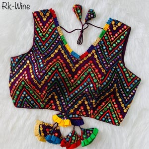 May include: A sleeveless, V-neck top in a deep purple hue, decorated with a vibrant chevron pattern of colourful beads and small mirrors. The neckline and edges are trimmed with rainbow-coloured accents. The top includes matching decorative tassels.