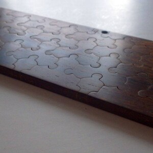 May include: A rectangular wooden puzzle with interlocking pieces. The puzzle is made of dark brown wood and has a smooth, polished finish. The puzzle pieces are cut in a classic jigsaw shape. The overall dimensions are approximately 12 inches long and 6 inches wide.
