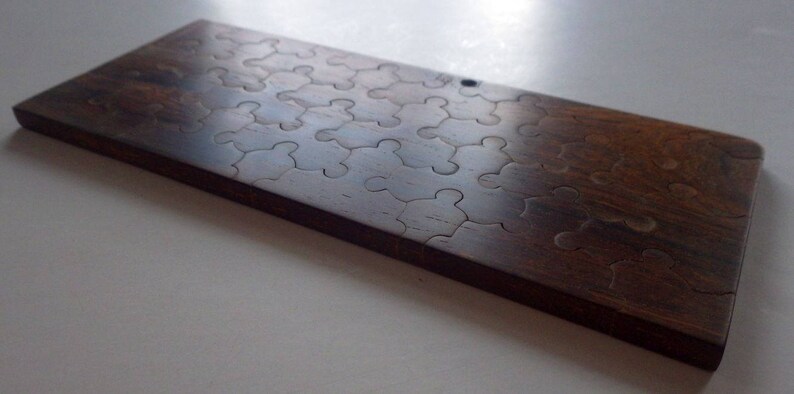 May include: A rectangular wooden puzzle with interlocking pieces. The puzzle is made of dark brown wood and has a smooth, polished finish. The puzzle pieces are cut in a classic jigsaw shape. The overall dimensions are approximately 12 inches long and 6 inches wide.