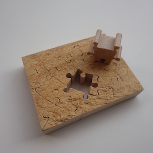 Pode incluir: Um quebra-cabeça de madeira com uma peça em falta e uma peça separada. O quebra-cabeça é feito de madeira marrom clara e tem formato quadrado. A peça em falta tem um formato único, e a peça separada é uma pequena estrutura tridimensional.