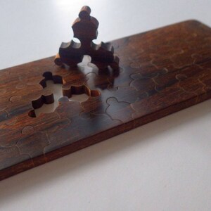 May include: A wooden puzzle with a dark brown finish. Several puzzle pieces are missing, with one piece partially inserted. The puzzle is rectangular and appears to be handmade. The wood grain is visible.