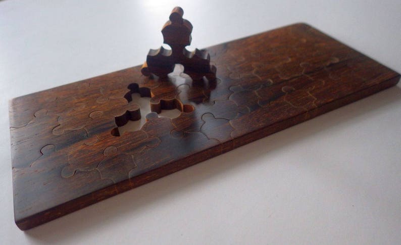 May include: A wooden puzzle with a dark brown finish. Several puzzle pieces are missing, with one piece partially inserted. The puzzle is rectangular and appears to be handmade. The wood grain is visible.