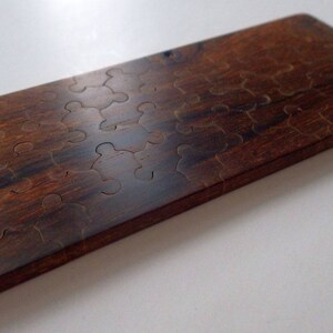 May include: A rectangular wooden puzzle with a dark brown finish. The puzzle pieces are cut into a unique shape, creating a challenging and engaging game. The wood grain is visible, adding to the natural aesthetic.