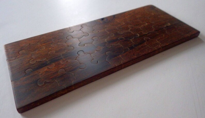 May include: A rectangular wooden puzzle with a dark brown finish. The puzzle pieces are cut into a unique shape, creating a challenging and engaging game. The wood grain is visible, adding to the natural aesthetic.