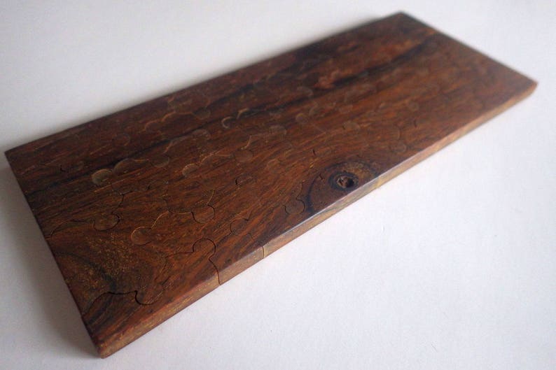 May include: A rectangular wooden puzzle with a rich, dark brown color and visible wood grain. The puzzle pieces are cut in a unique, organic shape. The wood has a smooth, polished finish, and the puzzle is set against a white background.