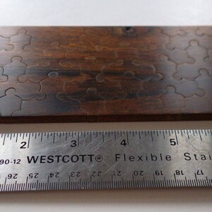 May include: A wooden jigsaw puzzle with interlocking pieces, stained in a dark brown color. The puzzle is rectangular and appears to be partially assembled. A metal ruler with inch measurements is placed below the puzzle.