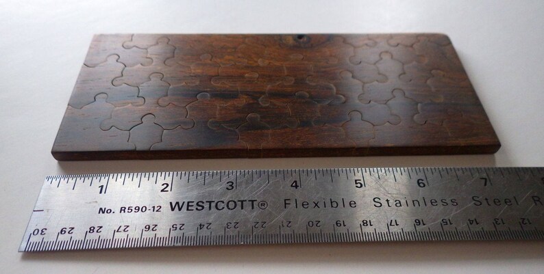 May include: A wooden jigsaw puzzle with interlocking pieces, stained in a dark brown color. The puzzle is rectangular and appears to be partially assembled. A metal ruler with inch measurements is placed below the puzzle.