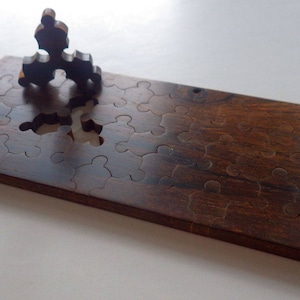May include: A wooden jigsaw puzzle with a dark brown finish. The puzzle is rectangular with several puzzle piece cutouts. One puzzle piece is partially inserted, and another is standing upright on the board. The puzzle is on a white surface.