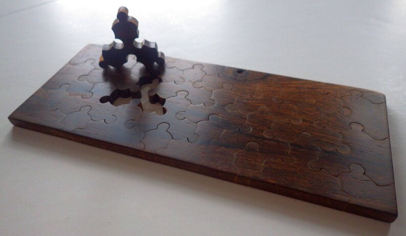May include: A wooden jigsaw puzzle with a dark brown finish. The puzzle is rectangular with several puzzle piece cutouts. One puzzle piece is partially inserted, and another is standing upright on the board. The puzzle is on a white surface.