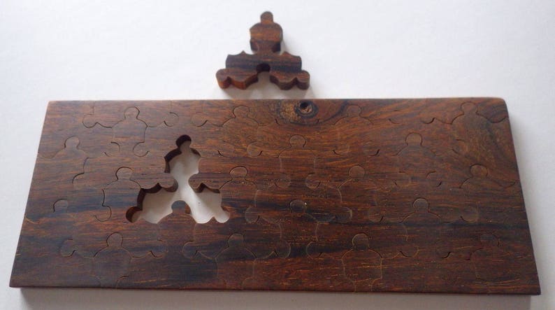 May include: A wooden jigsaw puzzle with a missing piece and a separate piece above. The puzzle is made of dark brown wood with visible grain. The puzzle pieces are shaped like people. The missing piece is in the center of the puzzle.