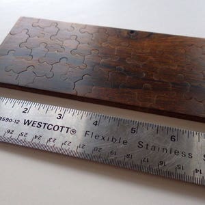 May include: A wooden puzzle with interlocking pieces, stained in a dark brown color. A stainless steel ruler, marked with inches, is placed next to the puzzle. The ruler is approximately 8 inches long.