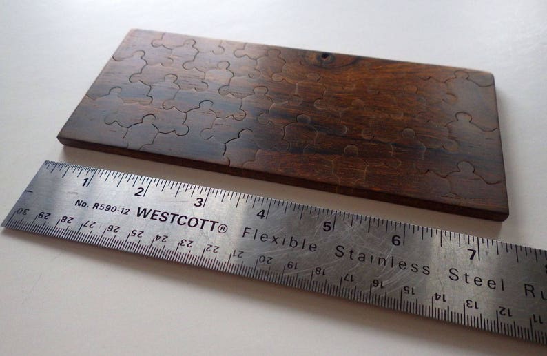 May include: A wooden puzzle with interlocking pieces, stained in a dark brown color. A stainless steel ruler, marked with inches, is placed next to the puzzle. The ruler is approximately 8 inches long.