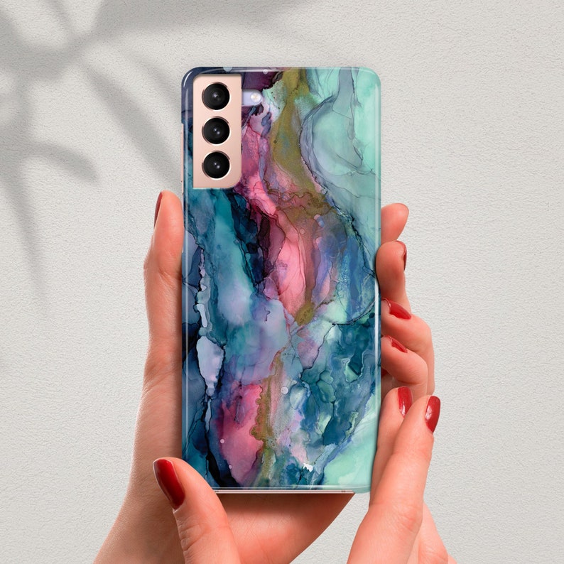 May include: A smartphone case with an abstract design in shades of teal, pink, and purple. The case has a glossy finish and a camera module with three lenses. The phone is held by a person with red nail polish.