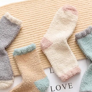 May include: Four pairs of fuzzy socks in different colors. The socks are made of a soft, fluffy material and have a contrasting color trim. The socks are arranged on a beige background.