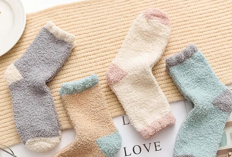 May include: Four pairs of fuzzy socks in different colors. The socks are made of a soft, fluffy material and have a contrasting color trim. The socks are arranged on a beige background.
